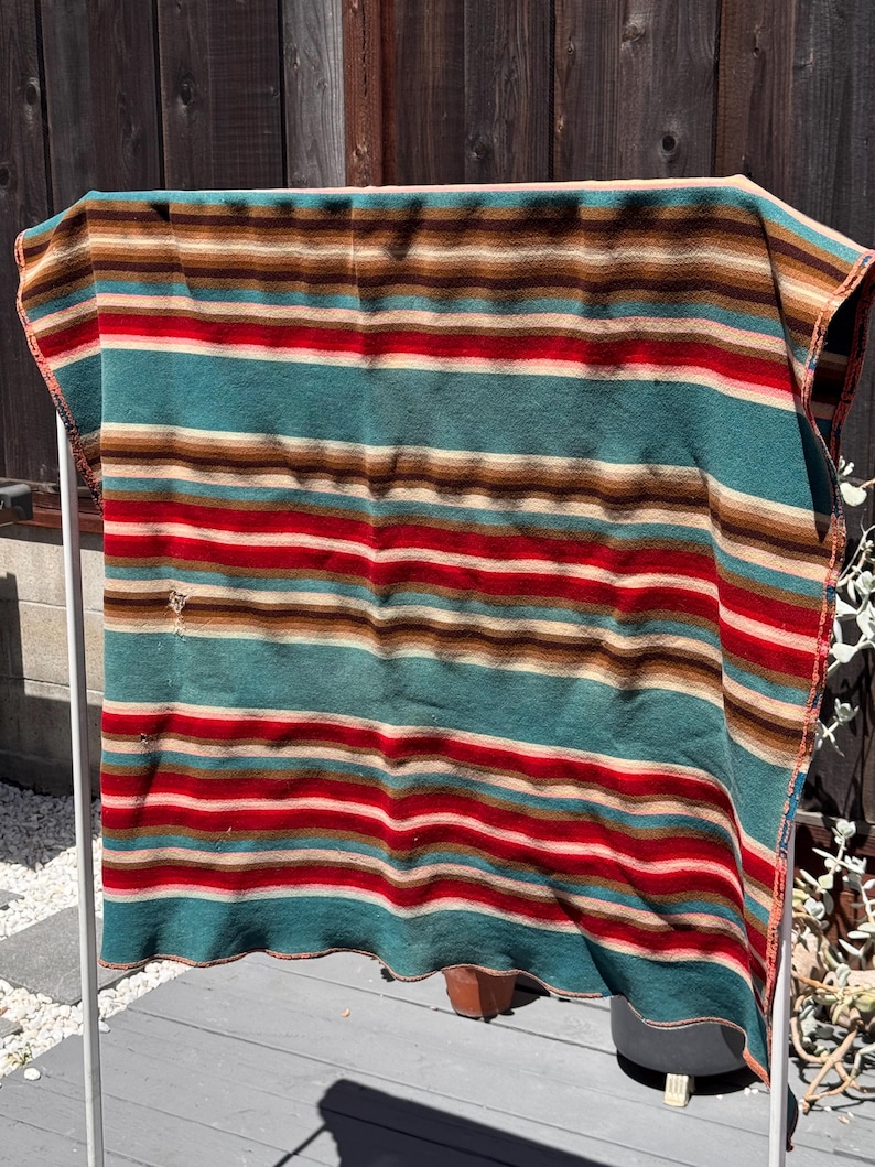 May include: A rectangular woven blanket featuring horizontal stripes in shades of teal, red, tan, and dark brown. The fabric displays a consistent pattern of varying widths across the entire surface, with a small visible tear on the left side.