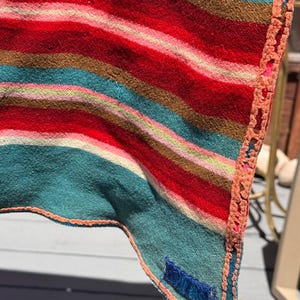 May include: A woven textile featuring horizontal stripes in shades of red, teal, brown, and pink. The fabric has a textured surface and is finished with a decorative orange crocheted border along the edge, including a small blue rectangular tag.