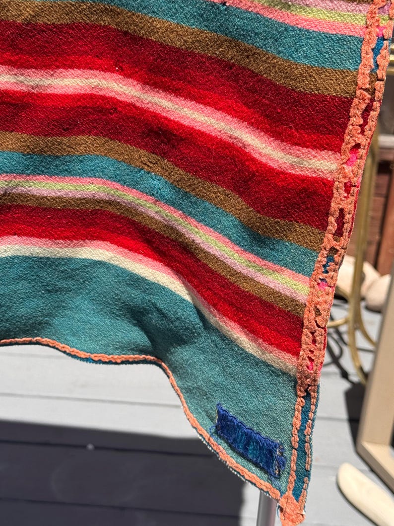 May include: A woven textile featuring horizontal stripes in shades of red, teal, brown, and pink. The fabric has a textured surface and is finished with a decorative orange crocheted border along the edge, including a small blue rectangular tag.