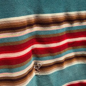 May include: A fabric garment featuring a horizontal striped pattern in shades of teal, brown, red, white, and light pink. The material displays a small, frayed hole located within the striped design, showing the texture of the woven textile.