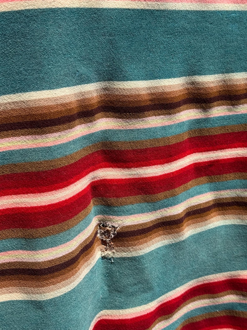 May include: A fabric garment featuring a horizontal striped pattern in shades of teal, brown, red, white, and light pink. The material displays a small, frayed hole located within the striped design, showing the texture of the woven textile.