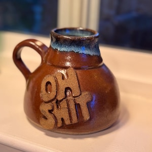 May include: A brown ceramic mug with a handle and the words "OH SHIT" embossed on the front. The rim of the mug has a blue glaze. The mug is sitting on a white surface.