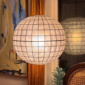 May include: A spherical pendant light fixture with a grid pattern. The light is white and the grid is black. The fixture is hanging from a black cord. The light is reflected in a window.