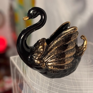 May include: A black ceramic swan figurine with gold accents on the wings and beak. The swan has a glossy finish and is posed with its neck curved. The figurine is likely a decorative item for a home.