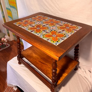 May include: A wooden side table with a rectangular top featuring a decorative tile inlay with orange and yellow floral patterns. The table has a lower shelf and turned legs, showcasing a warm, natural wood finish.