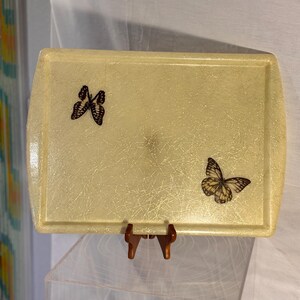 May include: A rectangular, pale yellow serving tray with a textured surface. Two butterfly decals are affixed to the tray. The tray is supported by a small, brown stand. The tray has rounded corners.