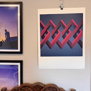 May include: A square art print featuring a repeating geometric pattern of interlocking red and dark maroon ribbons against a solid dark blue background. The print is suspended from a silver metal clip attached to a light-colored wall surface.