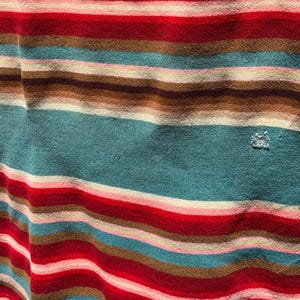 May include: A fabric featuring a horizontal striped pattern in shades of teal, red, brown, and light pink. The textile has a soft, slightly textured appearance with a small, visible snag or loose thread detail located on the right side of the fabric.
