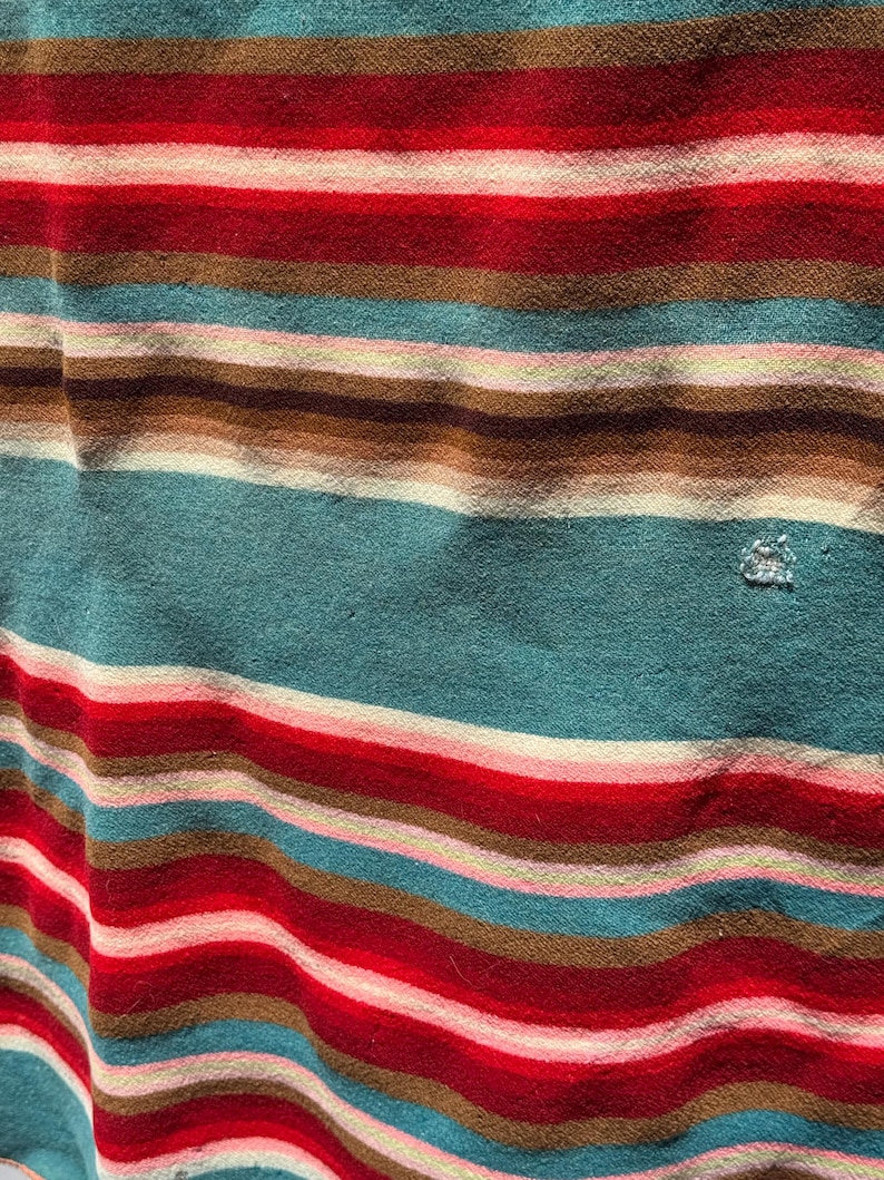 May include: A fabric featuring a horizontal striped pattern in shades of teal, red, brown, and light pink. The textile has a soft, slightly textured appearance with a small, visible snag or loose thread detail located on the right side of the fabric.