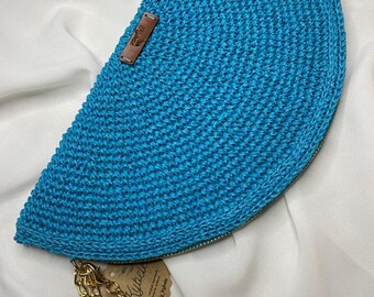 May include: A semi-circular bright blue crochet clutch bag featuring a textured stitch pattern, a gold-tone metal zipper closure, and a gold chain tassel. A small brown leather-like tag with the text Kybele is attached to the front surface.