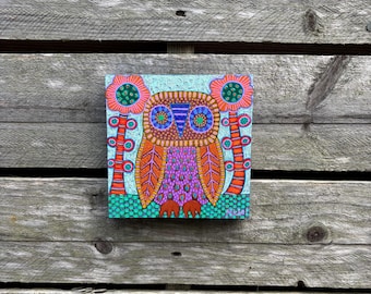May include: A colorful owl painting on a square canvas. The owl features orange, purple, and blue hues with intricate patterns. Two stylized red and green flowers flank the owl against a light blue background, set on a wooden plank backdrop.