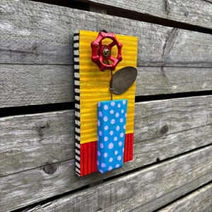 May include: A handmade wall art piece featuring a red valve handle flower with a metal leaf and a blue polka-dotted vase. The background is yellow with red and black stripes.