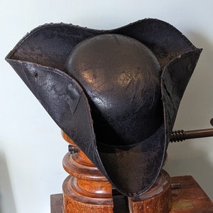 May include: A black leather tricorn hat with a wide brim and a tall crown. The hat is displayed on a wooden hat block.
