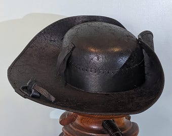 May include: A black leather tricorn hat with a wide brim, displayed on a wooden stand. The hat has a textured surface and is adorned with a small ribbon and buckle. A rolled parchment with a red ribbon sits nearby.