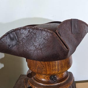 Pode incluir: Um chapéu tricornio de couro castanho, um estilo popular no século XVIII, é exibido em um suporte de chapéu de madeira. O chapéu tem uma superfície texturizada e é dobrado em três lados. O suporte do chapéu é feito de madeira polida.