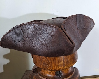 Può includere: Un cappello tricornio in pelle marrone, uno stile popolare nel XVIII secolo, è esposto su un espositore per cappelli in legno. Il cappello ha una superficie strutturata ed è ripiegato su tre lati. L'espositore per cappelli è in legno lucido.