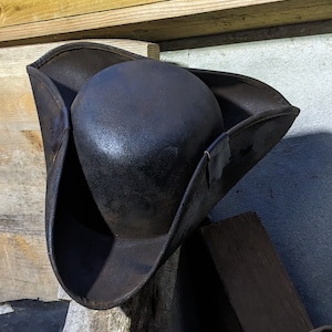Peut inclure: Un chapeau tricorne brun foncé et patiné, doté d'une calotte arrondie et d'un large bord retroussé sur trois côtés. Le chapeau présente une surface texturée et usée avec des signes visibles d'âge et d'utilisation, arborant une forme triangulaire.