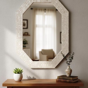 May include: An octagonal wall mirror with a mosaic-style, off-white frame hangs above a wooden console table. A small succulent in a white pot and a decorative vase with greenery sit on the table. Two books are stacked below the vase.