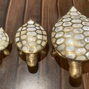 May include: Three brass turtle figurines with a mother of pearl shell design. The turtles are different sizes and are arranged in a row from largest to smallest.