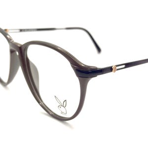 May include: Brown eyeglasses with a round frame and a thin, dark brown arm. The glasses feature a small gold-colored accent on the arm and a bunny logo on the lens.
