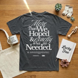 Op de afbeelding: Een grijs t-shirt met de tekst "More Than We Hoped & Exactly What We Needed." Een witte draagtas, een mok met de tekst "Tired as a Mother" en een notitieboek met de tekst "Lucky Few" zijn ook zichtbaar. De afbeelding bevat ook babyartikelen.