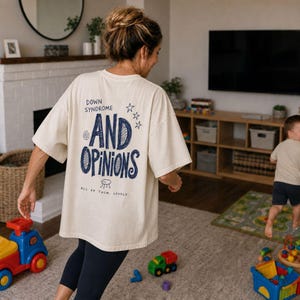 Op de afbeelding: Een oversized crèmekleurig T-shirt met donkerblauwe tekst op de achterkant die luidt Down Syndrome And Opinions All Of Them, Loudly, vergezeld van kleine ster- en spiraalafbeeldingen en een eenvoudige lijntekening van een klein wezen.