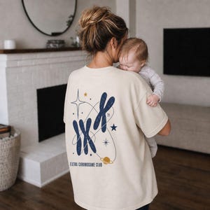 May include: A cream-colored t-shirt featuring a dark blue graphic on the back. The design includes three chromosome shapes, stars, a small planet, and the text "EXTRA CHROMOSOME CLUB" with the number 21 positioned near the chromosome structures.