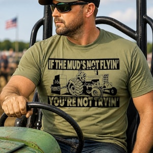 May include: An olive green short-sleeve t-shirt featuring a black graphic of a tractor pulling a weighted sled. The text reads IF THE MUD'S NOT FLYIN' and YOU'RE NOT TRYIN' with 16 TONS printed on the side of the sled in a bold font.