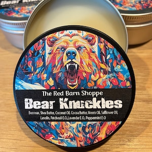 May include: A round tin container with a black lid featuring a colorful illustration of a bear's face. The lid has the text "The Red Barn Shoppe Bear Knuckles" and lists ingredients including beeswax, shea butter, coconut oil, cocoa butter, neem oil, safflower oil, lanolin, patchouli essential oil, lavender essential oil, and peppermint essential oil.