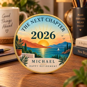 May include: A circular decorative plaque featuring a sunset landscape over water with mountains, a sailboat, and potted plants. The text reads THE NEXT CHAPTER, 2026, MICHAEL, and HAPPY RETIREMENT. The plaque has a gold-toned rim and a flat base.