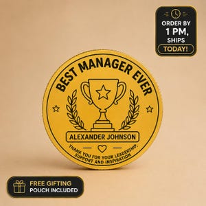Puede incluir: Un premio redondo de color dorado con el texto "BEST MANAGER EVER" y un gráfico de trofeo. El premio incluye el nombre "ALEXANDER JOHNSON" y un mensaje de agradecimiento. Una pequeña caja negra indica "FREE GIFTING POUCH INCLUDED".