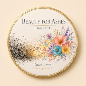Può includere: Un piatto decorativo rotondo color crema con bordo dorato. Il testo recita "BEAUTY FOR ASHES", "Isaiah 61:3" e "Grace 2026". Una composizione floreale nei toni del rosa, blu e viola è affiancata da un motivo maculato nero e oro.