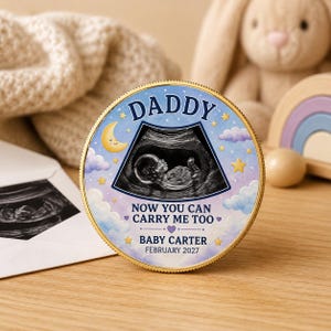 May include: A round gold-rimmed coin featuring a central ultrasound image of a fetus. The design includes a yellow crescent moon, purple clouds, and gold stars on a light blue background. Text reads: DADDY NOW YOU CAN CARRY ME TOO BABY CARTER FEBRUARY 2027.