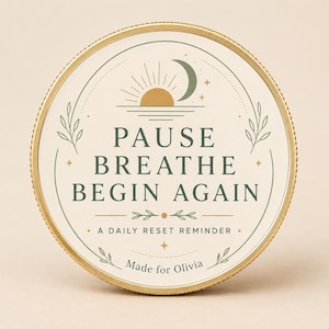 May include: A round gold-rimmed token featuring a cream-colored face with the text PAUSE BREATHE BEGIN AGAIN. The design includes a gold sun, a green crescent moon, green leaf accents, and the text A DAILY RESET REMINDER and MADE FOR OLIVIA.