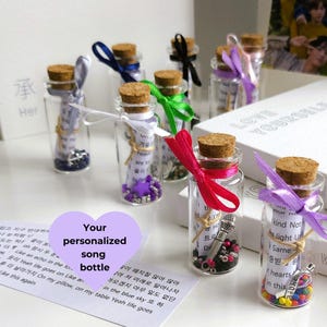 Könnte beinhalten: Eine Sammlung kleiner Glasflaschen mit Korkstopfen, gefüllt mit bunten Perlen und aufgerollte Papier. Die Flaschen sind mit Bändern verziert und auf einer weißen Oberfläche angeordnet. Der Text "Your personalized song bottle" ist auf einem violetten Aufkleber geschrieben. Die Flaschen sind von Büchern mit dem Text "Love Yourself" auf dem Cover umgeben.