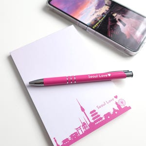 May include: A pink pen with the text "Seoul Love" rests on a white notepad with a pink Seoul skyline graphic. A smartphone with a clear case is in the background. The pen and notepad are on a white surface.