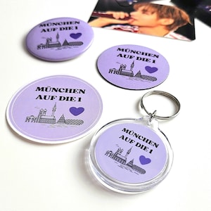 May include: Two circular lavender badges, one lavender sticker, and one clear acrylic keyring. Each item features the black text MÜNCHEN AUF DIE 1, a black line drawing of the Munich skyline, and a solid purple heart icon on a light purple background.