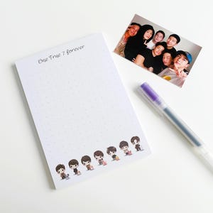 Könnte beinhalten: Ein weißer Notizblock mit einem gepunkteten Muster und dem Text "One True 7 forever" oben. Der Notizblock hat unten kleine Comiczeichnungen von sieben Personen. Ein violetter Stift liegt auf einer weißen Oberfläche neben dem Notizblock.