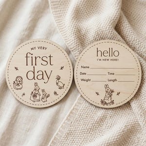 May include: Two round wooden baby milestone discs. One says "MY VERY first day" with illustrations of animals. The other says "hello I'M NEW HERE!" with fill-in-the-blank fields for baby's information. The discs are on a cream-colored blanket.