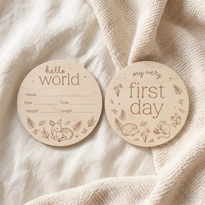 Può includere: Due targhe circolari in legno chiaro con testo e illustrazioni incise in marrone. Una targa riporta hello world con righe per nome, data, ora, peso e lunghezza, con un cerbiatto. L'altra riporta my very first day con un riccio e un topolino.