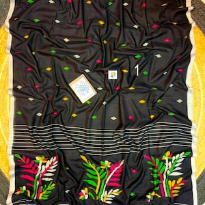 May include: A black textile featuring a pattern of small, scattered diamond-shaped motifs in yellow, green, and pink. The lower section displays horizontal white stripes and three large, colorful floral designs in pink, green, and yellow.