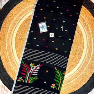 May include: A black textile featuring a pattern of small green, yellow, and pink diamond shapes, horizontal white stripes, and large floral motifs in pink, green, and white. A small tag displays the number 1 and a logo with the text Handloom.