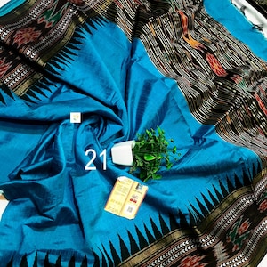 May include: A bright blue saree featuring a wide black border with intricate ikat patterns in red, white, and green. The fabric has a subtle sheen and a prominent black triangular motif along the edge. A small white tag with the number 21 is visible.