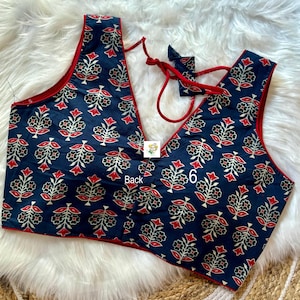 May include: A navy blue sleeveless top with a V-neck and a floral pattern in red, white, and black. The edges are trimmed with red fabric. The word "Back" is visible on the garment. The top has a tie closure.