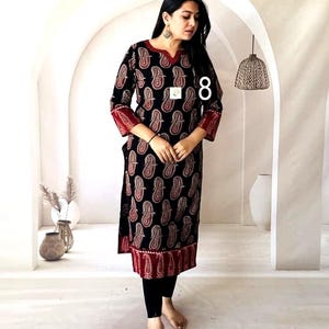 Puede incluir: Vestido t&uacute;nica negro con estampado de cachemira en rojo y blanco. El vestido tiene mangas tres cuartos con pu&ntilde;os rojos y un borde rojo en el dobladillo. La modelo lleva leggings negros. El n&uacute;mero "8" es visible en el vestido.