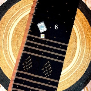 May include: A black saree featuring a copper-colored border and horizontal stripes. The fabric is decorated with small gold-toned dots and two diamond-shaped geometric patterns. A small white tag with the number 6 and a logo is attached.