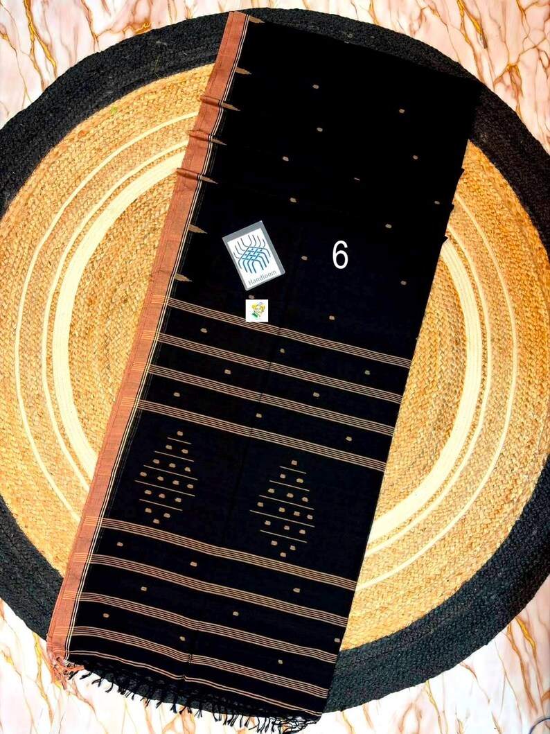 May include: A black saree featuring a copper-colored border and horizontal stripes. The fabric is decorated with small gold-toned dots and two diamond-shaped geometric patterns. A small white tag with the number 6 and a logo is attached.