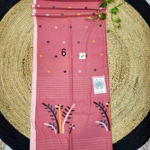 May include: A dusty rose rectangular textile featuring a pattern of small, multicolored geometric shapes. The lower section displays a stylized floral motif in muted tones, horizontal white stripes, and a fringed edge. A label reads Handloom.