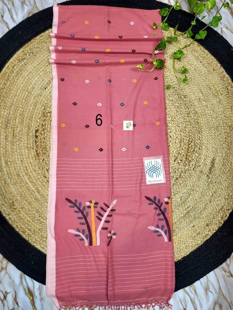 May include: A dusty rose rectangular textile featuring a pattern of small, multicolored geometric shapes. The lower section displays a stylized floral motif in muted tones, horizontal white stripes, and a fringed edge. A label reads Handloom.