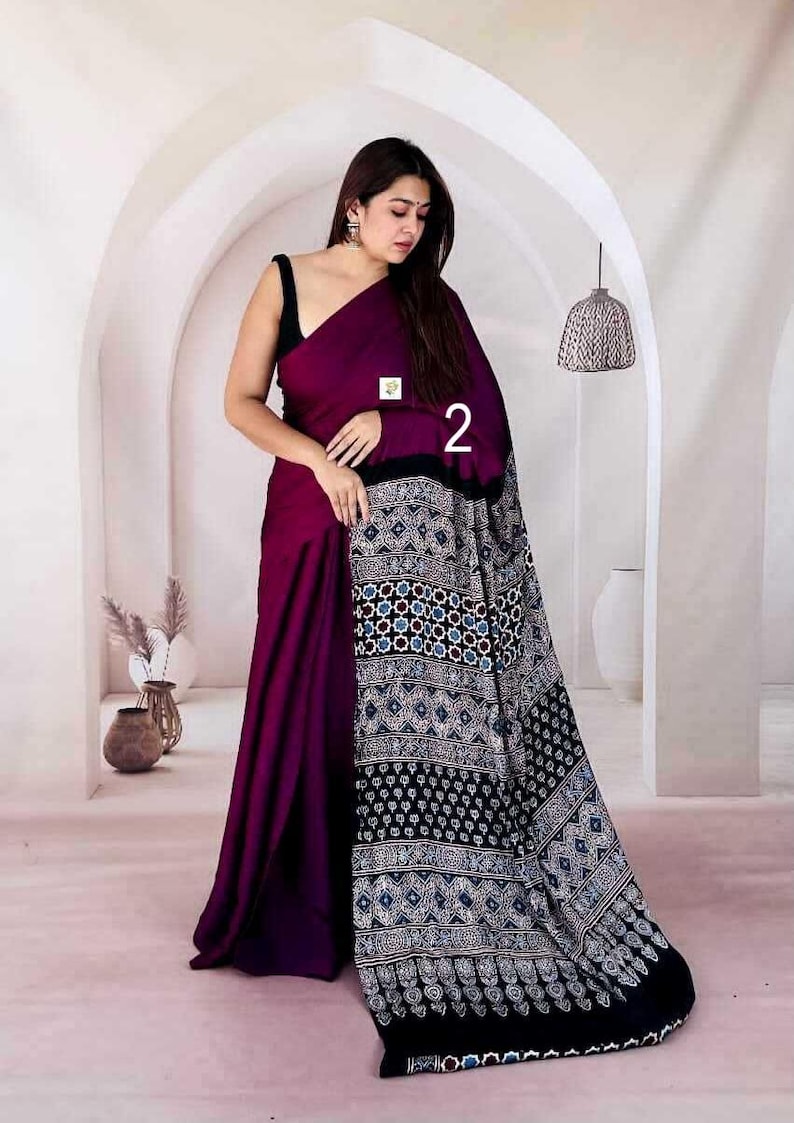 Puede incluir: Un sari de color morado intenso con un cuerpo de color s&oacute;lido y un pallu negro en contraste adornado con intrincados patrones geom&eacute;tricos en blanco, azul y negro. La prenda incluye el n&uacute;mero 2 impreso y una peque&ntilde;a etiqueta cuadrada.